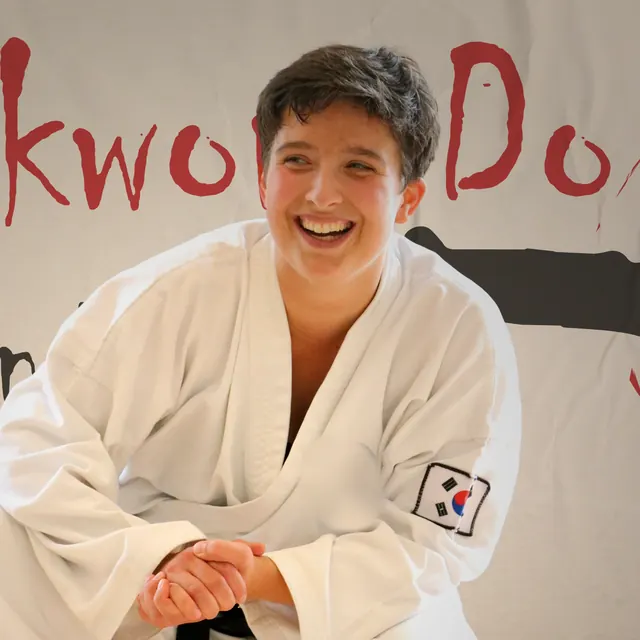 Jenny Steinberger im weißen Taekwon-Do-Anzug, lachend und leicht vorgebeugt mit gefalteten Händen, hinter ihr ein Banner mit dem roten Schriftzug Taekwon-Do