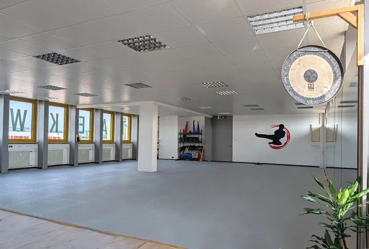 Heller, leerer Trainingsraum mit grauen Matten, großen Fenstern mit dem Schriftzug TAEKWONDO, einem Gong und dem Logo der Schule an der Wand