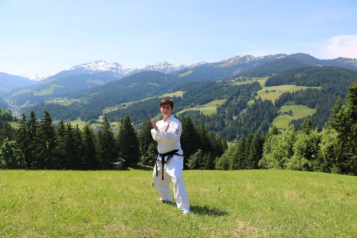 Schwarzgurt im weißen Taekwon-Do-Anzug übt eine Form auf einer Bergwiese, dahinter Alpengipfel und Wald