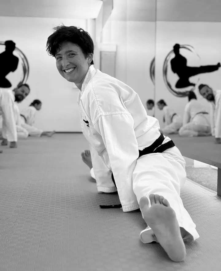 Jenny Steinberger im weißen Taekwon-Do-Anzug mit schwarzem Gürtel im Spagat auf der Matte, lacht in die Kamera, im Hintergrund dehnen sich weitere Schüler vor der Spiegelwand mit dem Logo der Schule