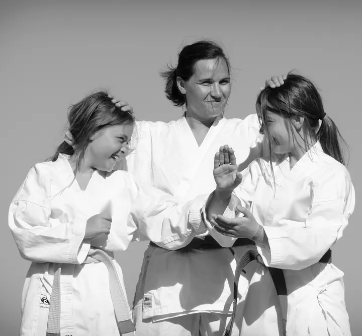 Eine Frau im weißen Taekwon-Do-Anzug steht lachend zwischen zwei Mädchen mit gelbem und grünem Gürtel und legt beiden eine Hand auf den Kopf, die Mädchen lachen sich an