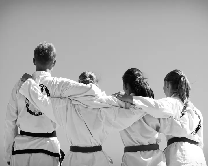 Vier Jugendliche im weißen Taekwon-Do-Anzug mit verschiedenen Gürtelfarben stehen von hinten gesehen Arm in Arm nebeneinander