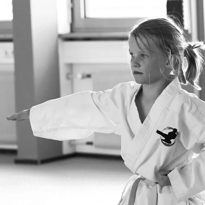 Junge im weißen Taekwon-Do-Anzug mit weißem Gürtel, ein Arm waagerecht zur Seite gestreckt, die andere Hand an der Hüfte, Blick konzentriert nach vorn