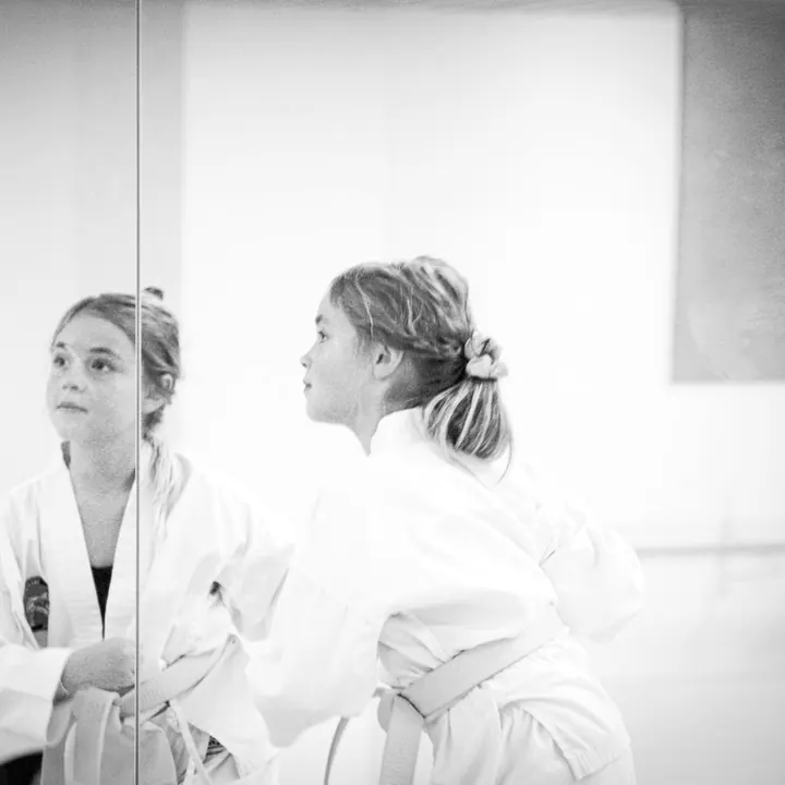 Mädchen im weißen Taekwon-Do-Anzug mit gelbem Gürtel steht konzentriert im Profil vor der Spiegelwand, links ihr Spiegelbild