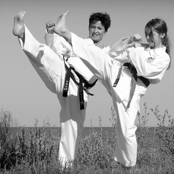 Eine Frau mit schwarzem Gürtel und ein Mädchen mit grünem Gürtel, beide im weißen Taekwon-Do-Anzug, machen draußen vor dem Meer gleichzeitig einen hohen Kick