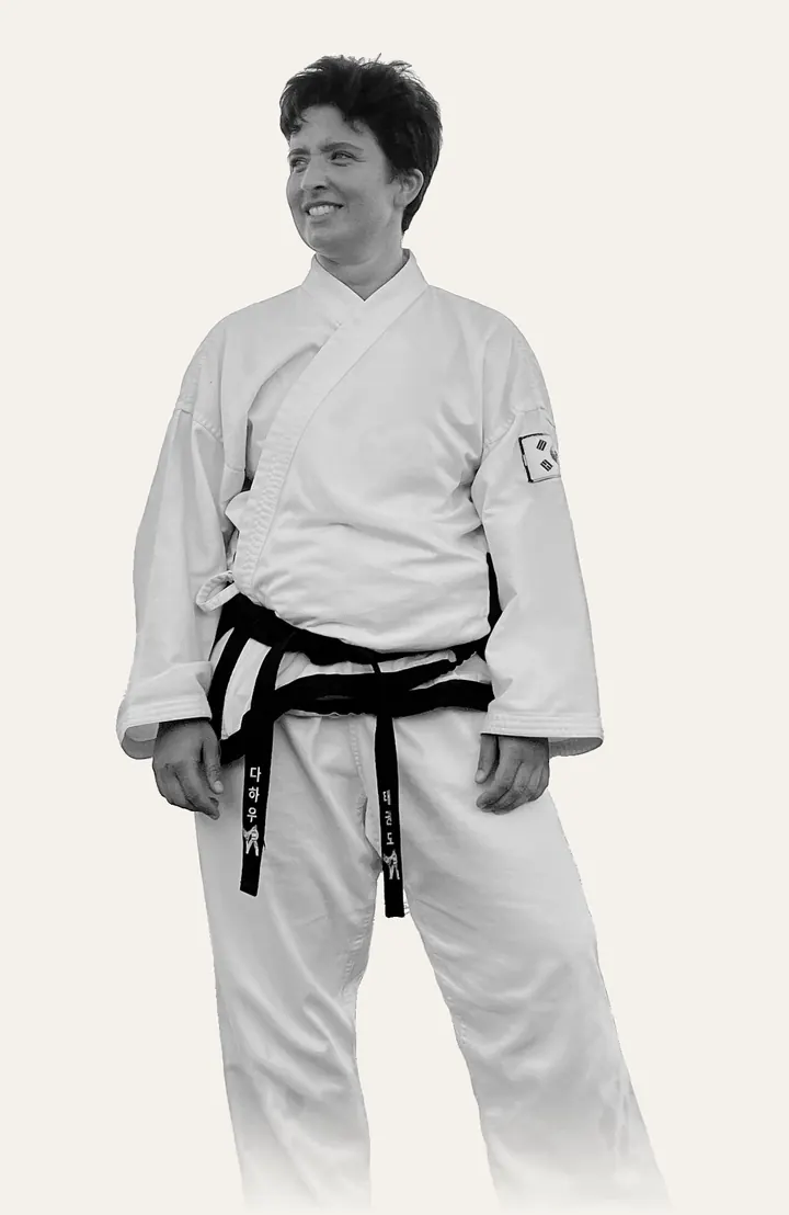 Schulleiterin Jenny Steinberger im weißen Taekwon-Do-Anzug mit schwarzem Gürtel und koreanischer Flagge am Ärmel, steht entspannt und lächelt zur Seite