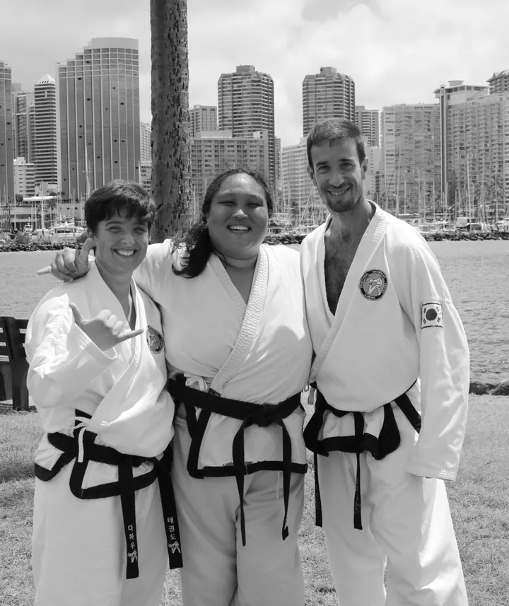 Drei Schwarzgurte im weißen Taekwon-Do-Anzug, Arm in Arm und lachend, vor Hochhäusern und einem Hafen im Trainingslager