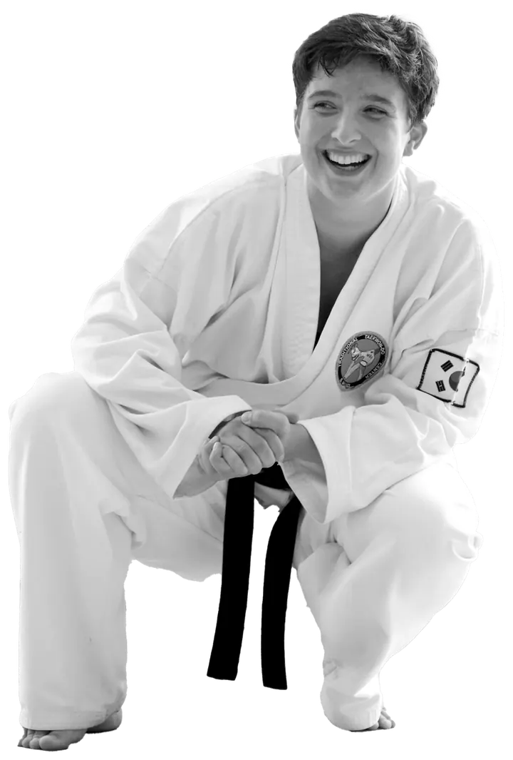 Jenny Steinberger im weißen Taekwon-Do-Anzug mit schwarzem Gürtel, in der Hocke mit gefalteten Händen, lacht herzlich